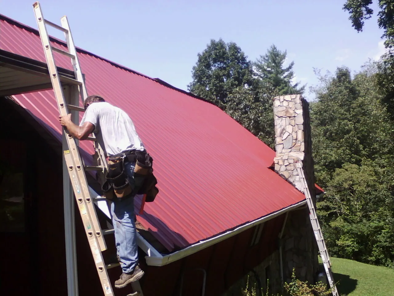 Roofer on a ladder beside a red metal roof and stone chimney — Soffit & Fascia Repair by Roofing Brothers in Adamsville
